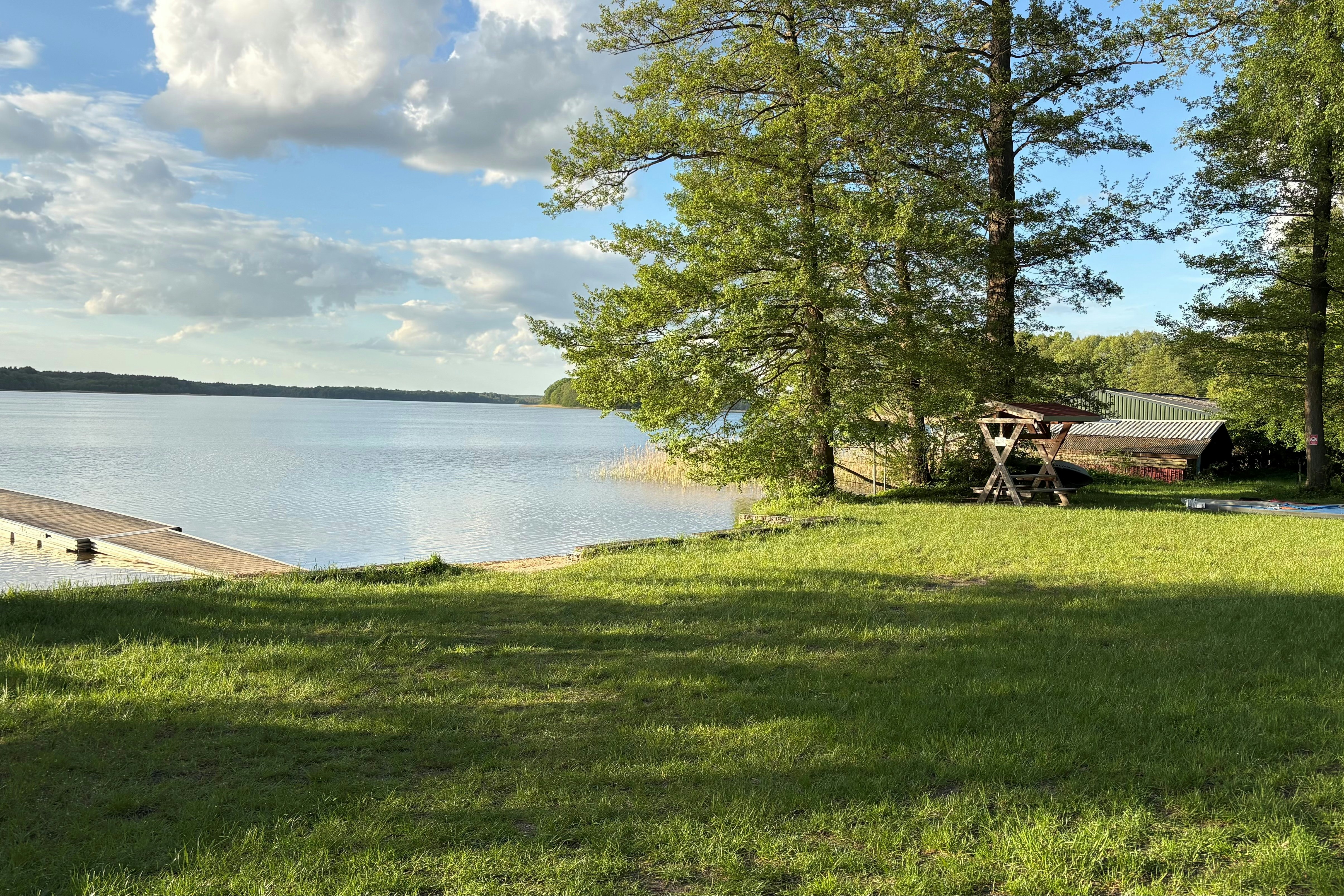 Grüner Campingplatz am See mit Uferpromenade, Bäumen und Sitzgelegenheiten am Rand des Wassers.