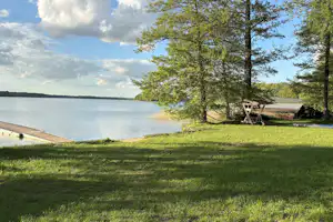Grüner Campingplatz am See mit Uferpromenade, Bäumen und Sitzgelegenheiten am Rand des Wassers.
