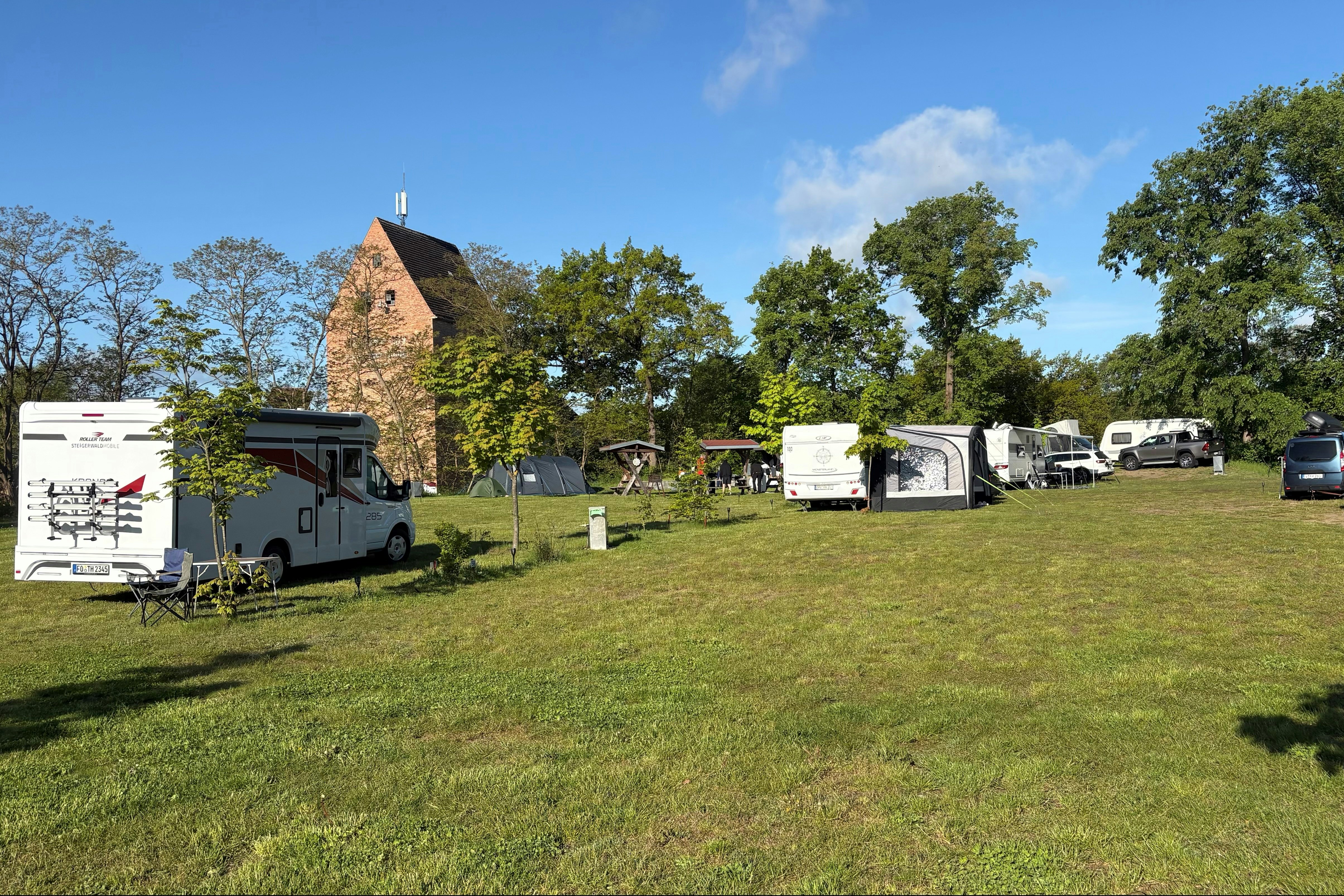 Campingplatz mit Wohnwagen und Autos auf einer grünen Wiese, im Hintergrund ein Backsteingebäude und Bäume.