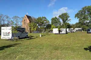 Campingplatz mit Wohnwagen und Autos auf einer grünen Wiese, im Hintergrund ein Backsteingebäude und Bäume.