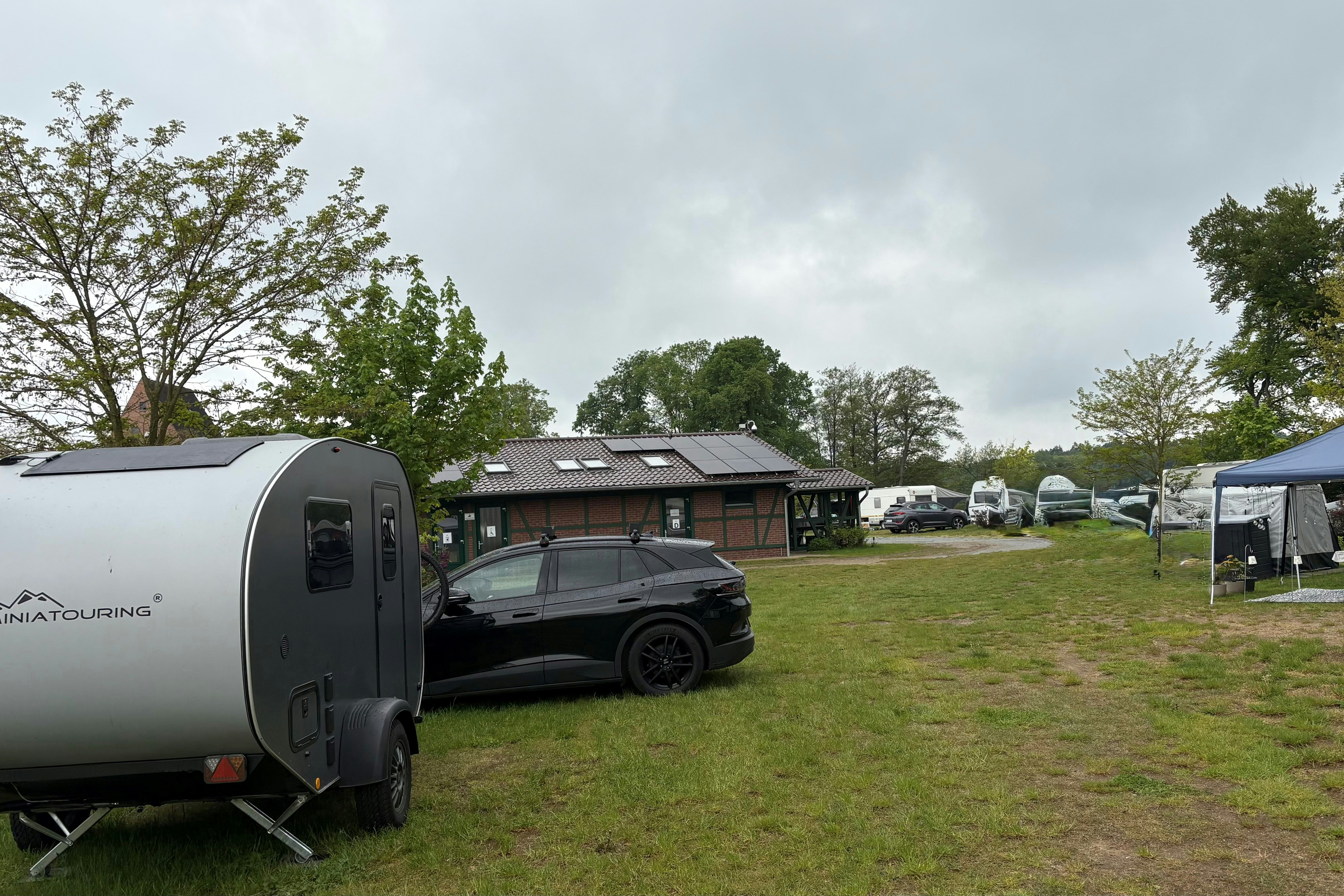 Wohnwagen-Anhänger auf einem Campingplatz, daneben Auto und Zelt auf der Wiese, im Hintergrund weitere Stellplätze und Bäume.