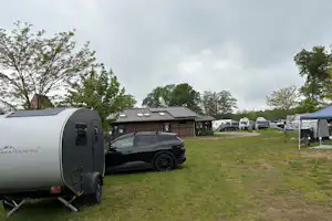 Wohnwagen-Anhänger auf einem Campingplatz, daneben Auto und Zelt auf der Wiese, im Hintergrund weitere Stellplätze und Bäume.