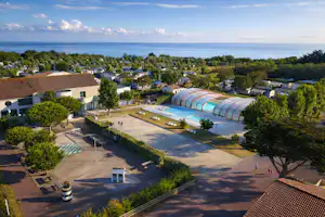 Campinganlage mit überdachtem Schwimmbad, Wohngebäuden und Wegen, dahinter das Meer und Bäume im Hintergrund.