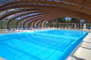 Überdachtes Schwimmbecken mit blauem Wasser, Liegen am Beckenrand und Glasfront im Hintergrund.