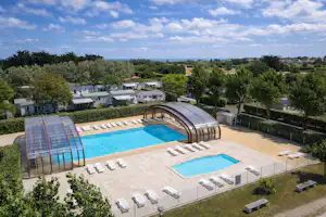 Überdachtes Schwimmbecken und weiteres Becken auf dem Campingplatz, umgeben von Liegen, Wegen und Bäumen.