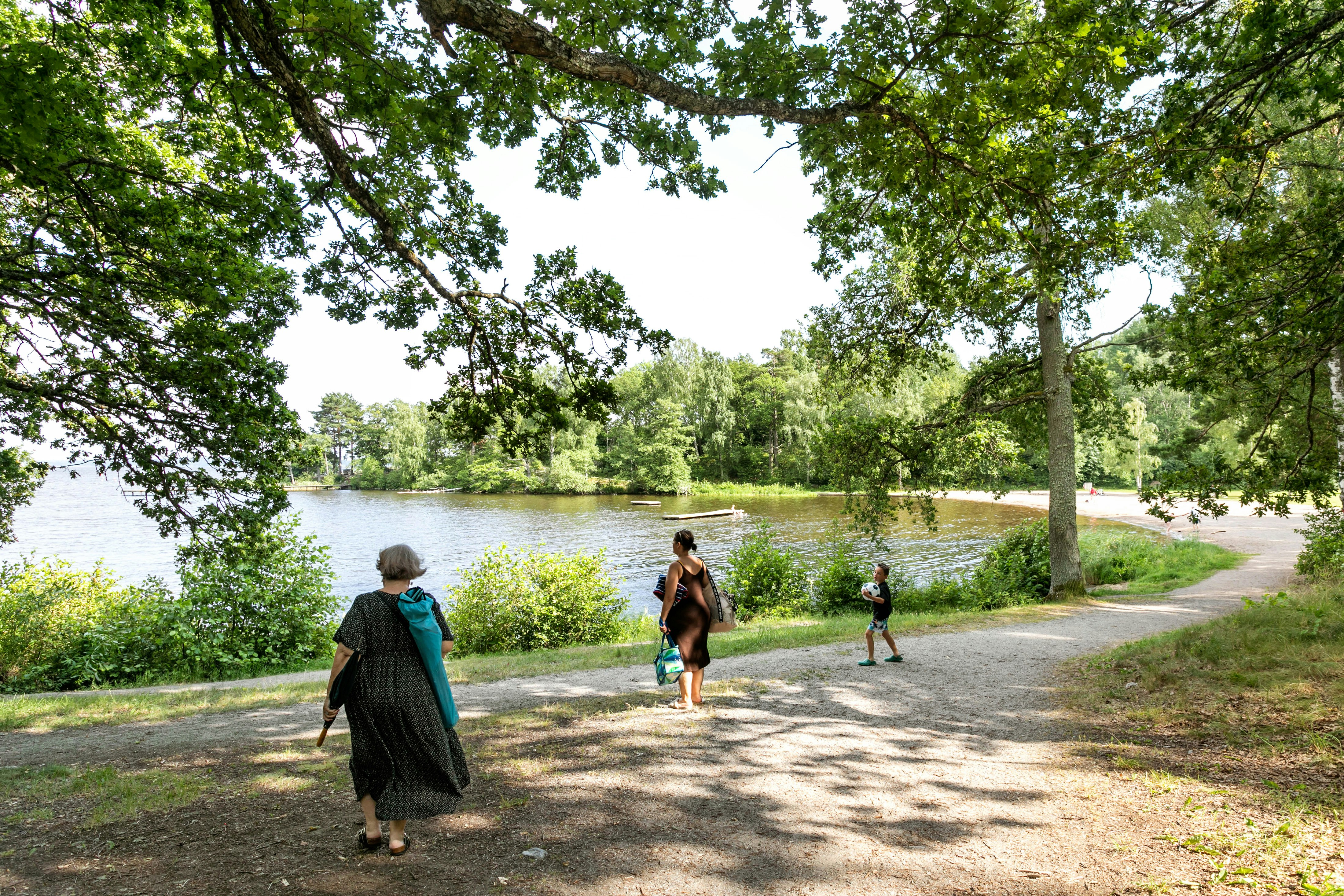 Familie läuft auf einem Weg am See entlang, umgeben von Bäumen und Grün, im Hintergrund das ruhige Wasser.