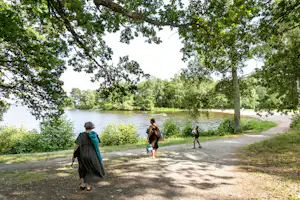 Familie läuft auf einem Weg am See entlang, umgeben von Bäumen und Grün, im Hintergrund das ruhige Wasser.