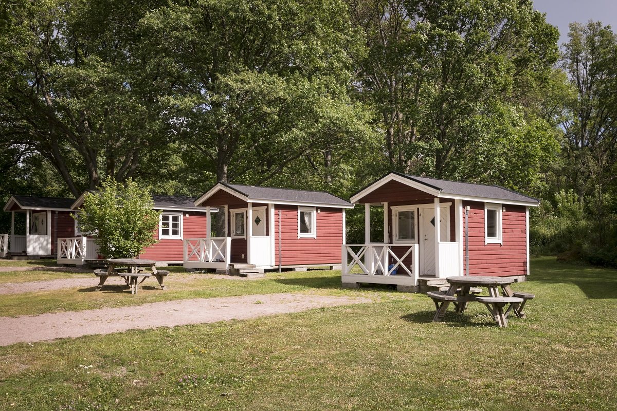 Rote Holz-Cabins mit Veranda, Picknicktischen und Sitzbänken auf einem gepflegten Campingplatz, umgeben von Bäumen.