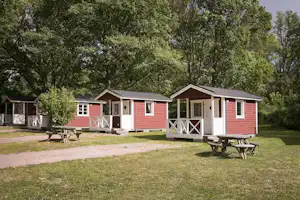 Rote Holz-Cabins mit Veranda, Picknicktischen und Sitzbänken auf einem gepflegten Campingplatz, umgeben von Bäumen.