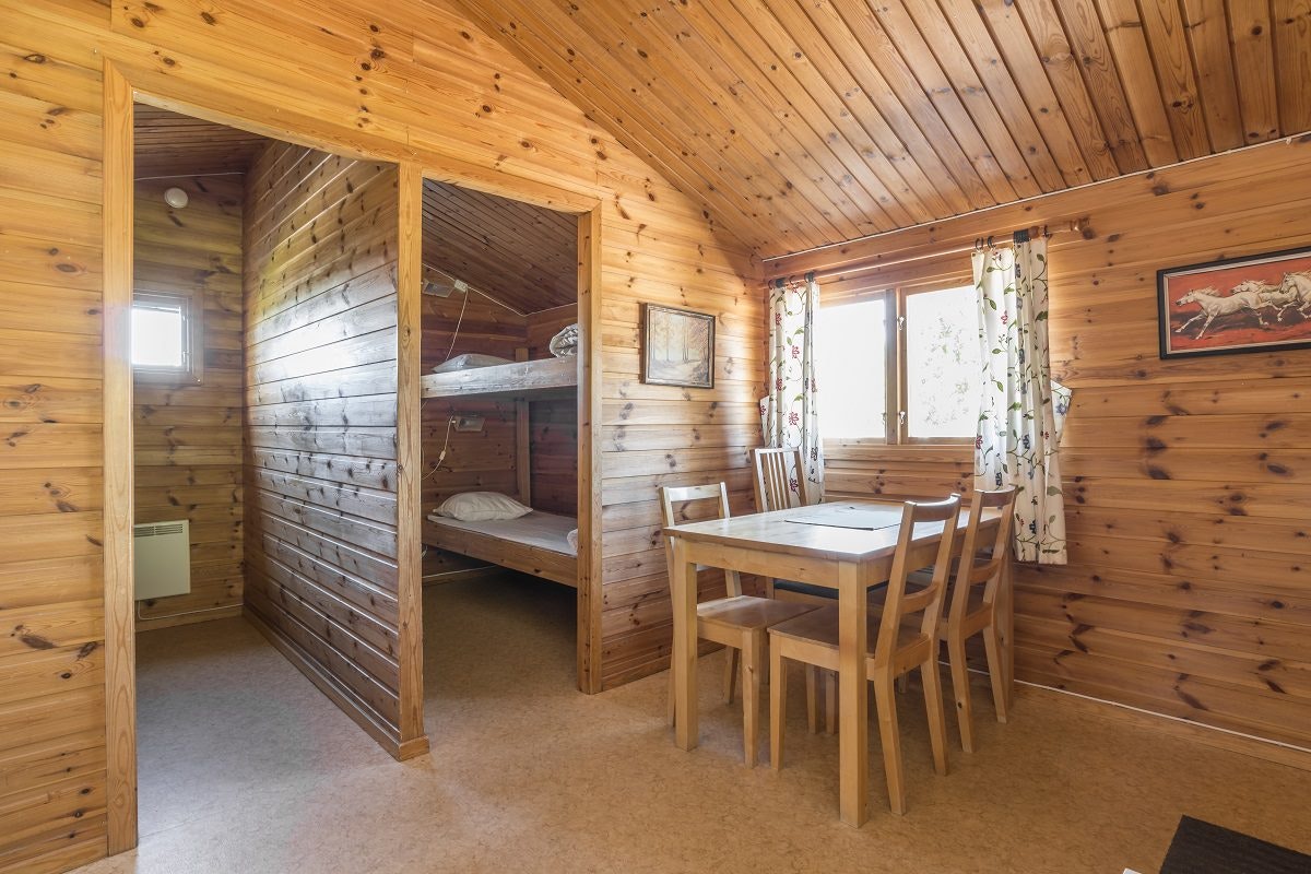 Innenansicht eines Holzhauses mit Esstisch und Stühlen, Vorhang am Fenster und Schlafbereich mit Etagenbett.