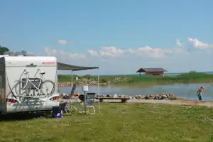 Wohnwagen am See mit Fahrrad am Heck, Sonnendach und Sitzbereich am Ufer, im Hintergrund Steg und Schilf.