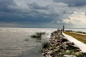 Seeufer am Campingplatz: Betonweg, Steinhaufen und Sitzgruppe am Wasser unter bewölktem Himmel.