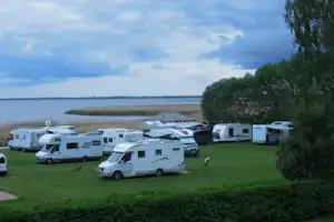 Reihe von Wohnmobilen auf einem grünen Campingplatz am See, im Hintergrund Wasser und Bäume.
