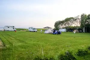 Campingplatz mit Wohnmobilen und Zelt auf grünem Rasen, Bäume am Rand und Blick auf das Wasser in der Ferne.