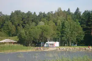Wohnwagen und Camper auf einem Campingplatz am Rand eines Sees, umgeben von Bäumen und einem Kiesweg.