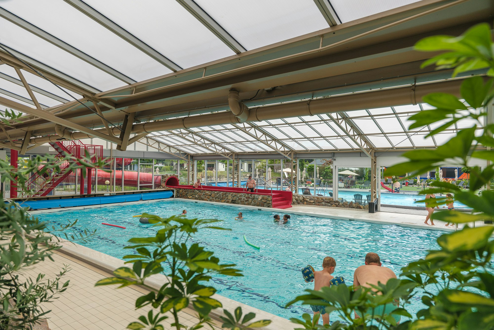 Innenpool mit roter Rutsche, Überdachung aus Glas und Stahlträgern, Kinder planschen im Wasser.