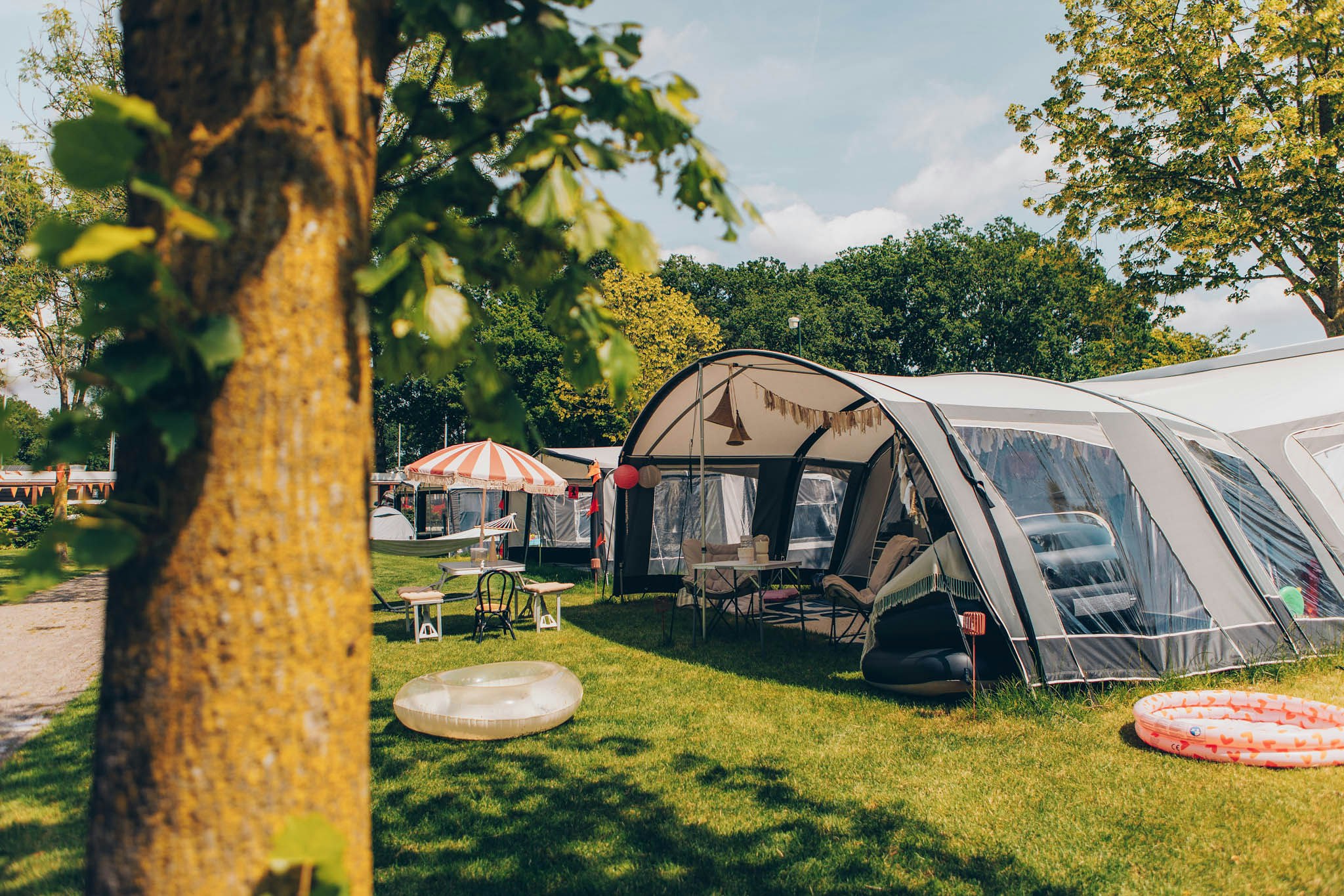 Campingzelte und Wohnwagen auf einem grünen Stellplatz, mit Picknicktisch und Liege auf dem Rasen, Bäume im Vordergrund.
