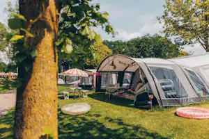 Campingzelte und Wohnwagen auf einem grünen Stellplatz, mit Picknicktisch und Liege auf dem Rasen, Bäume im Vordergrund.
