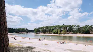 Sandstrand am See mit vielen Liegen und Paddelbooten, im Hintergrund Bäume und Uferlinie.