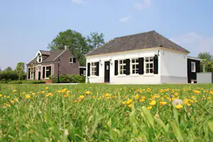 Weiße Ferienhaus-Terrasse mit dunklen Fenstern vor einem gepflegten Garten, im Vordergrund blühende Wiese.