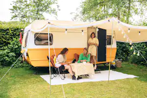Wohnwagen-Zelt mit Terrasse, Tisch und Stühlen, Lichterkette über dem Eingang, umgeben von Büschen und Bäumen.