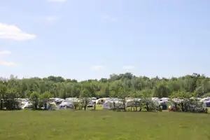 Wohnwagen und Zelte auf einem Campingplatz, davor eine große Wiese, im Hintergrund Bäume und ein Weg.
