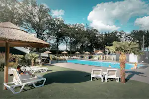 Poolbereich auf einem Campingplatz mit Liegen, Sonnenschirmen, Palmen und Bäumen, im Hintergrund weitere Unterkünfte.
