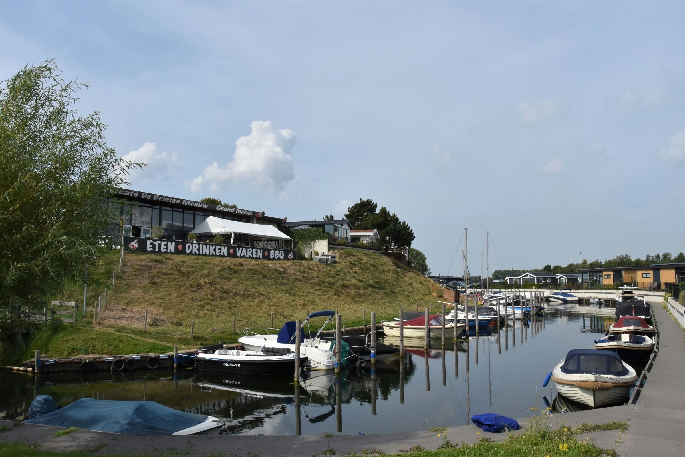 Hafen mit Stegen und Booten am ruhigen Wasser, im Hintergrund Gebäude und ein Schild an der Uferböschung.
