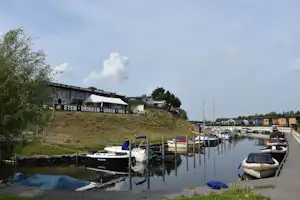 Hafen mit Stegen und Booten am ruhigen Wasser, im Hintergrund Gebäude und ein Schild an der Uferböschung.