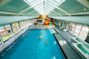 Innenpool mit blauem Wasser, großen Fenstern und Glasdach, im Hintergrund Spielbereich mit Rutsche und Figuren.