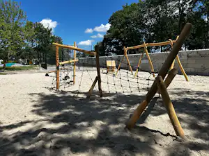 Klettergerüst aus Holz mit Seilnetz auf Sand, daneben Schaukel und Bänke, im Hintergrund Zaun und Bäume.