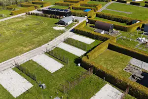 Campingstellplätze auf Rasen mit gepflasterten Flächen, Hecken und Wegen, im Hintergrund ein Gebäude mit Solarpaneln.