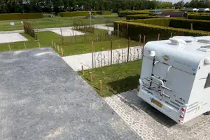 Wohnwagen auf einem Stellplatz mit gepflastertem Weg, Hecken und Zäunen, im Hintergrund Rasen und weitere Fahrzeuge.