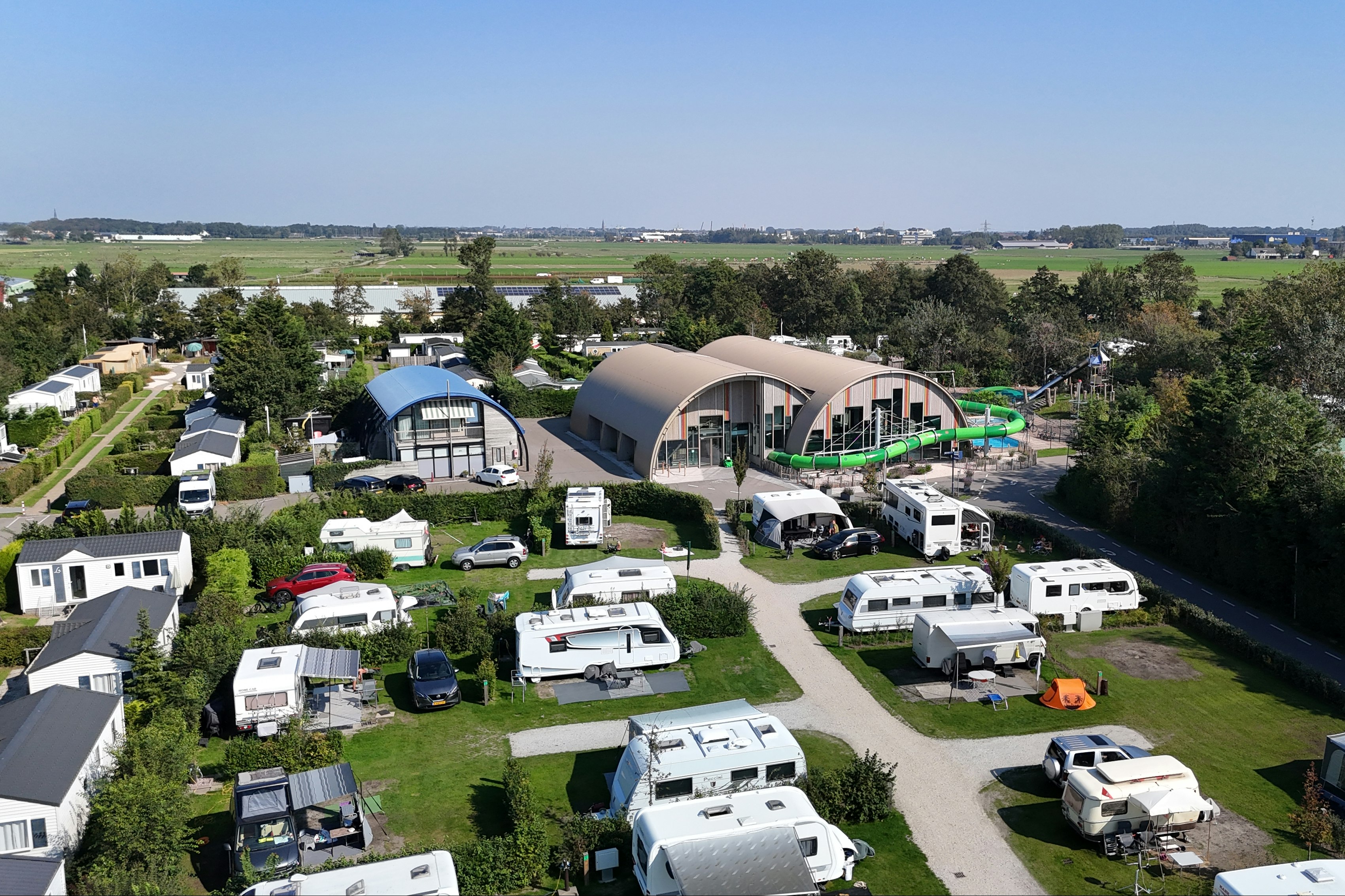 Luftaufnahme eines Campingplatzes mit Wohnmobilen und Wohnwagen, gepflegten Stellplätzen, Wegen und einem großen Gemeinschaftsgebäude mit Glasfront.