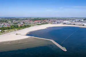 Luftaufnahme einer Bucht mit Sandstrand, Hafenmole und Wasser, dahinter eine Stadt mit Gebäuden und Straßen.