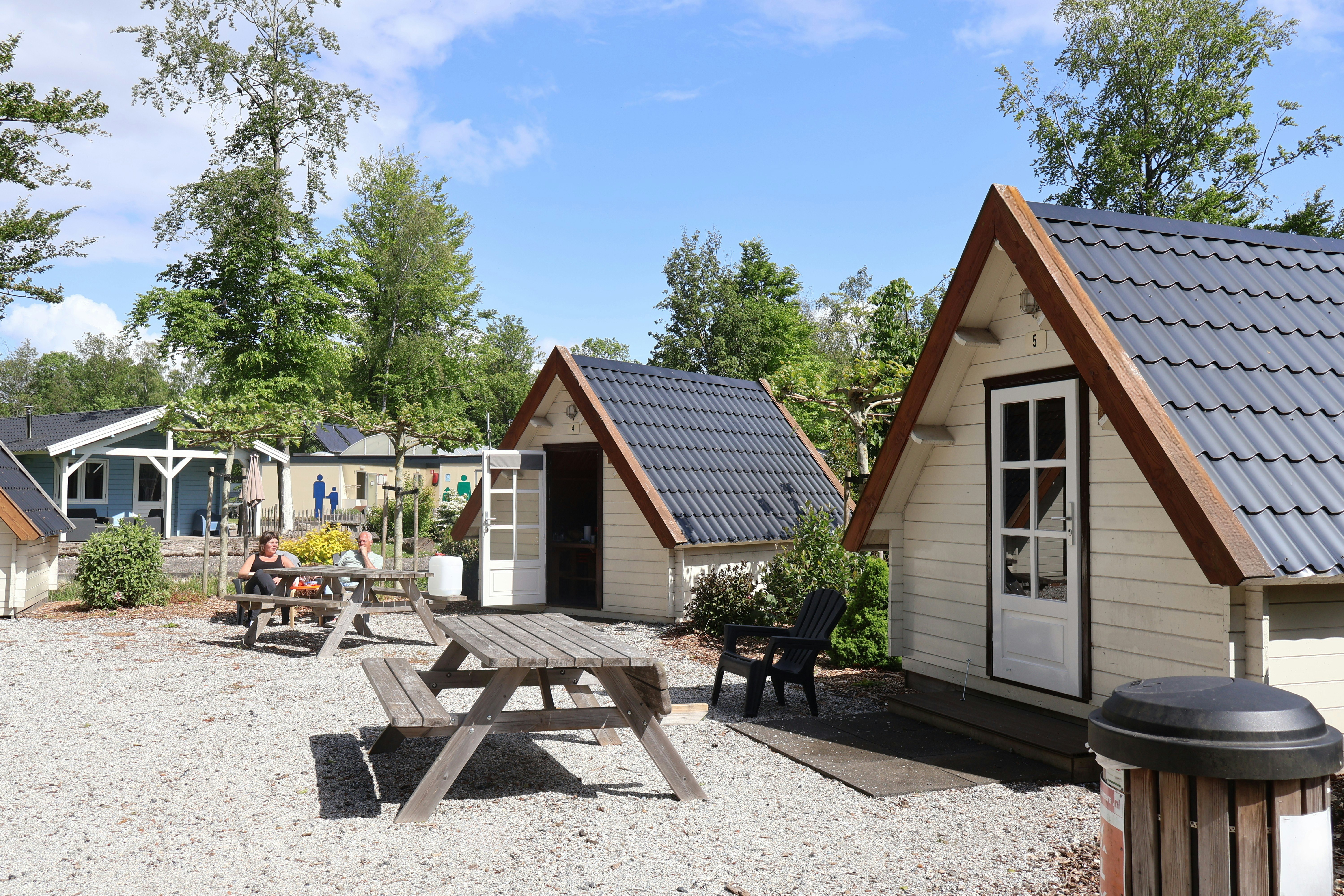 Holzhäuser und Zelteinheiten auf einem Campingplatz, mit Terrasse, Tisch und Stühlen sowie Grill am Rand, umgeben von Bäumen.