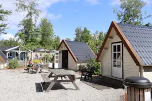 Holzhäuser und Zelteinheiten auf einem Campingplatz, mit Terrasse, Tisch und Stühlen sowie Grill am Rand, umgeben von Bäumen.