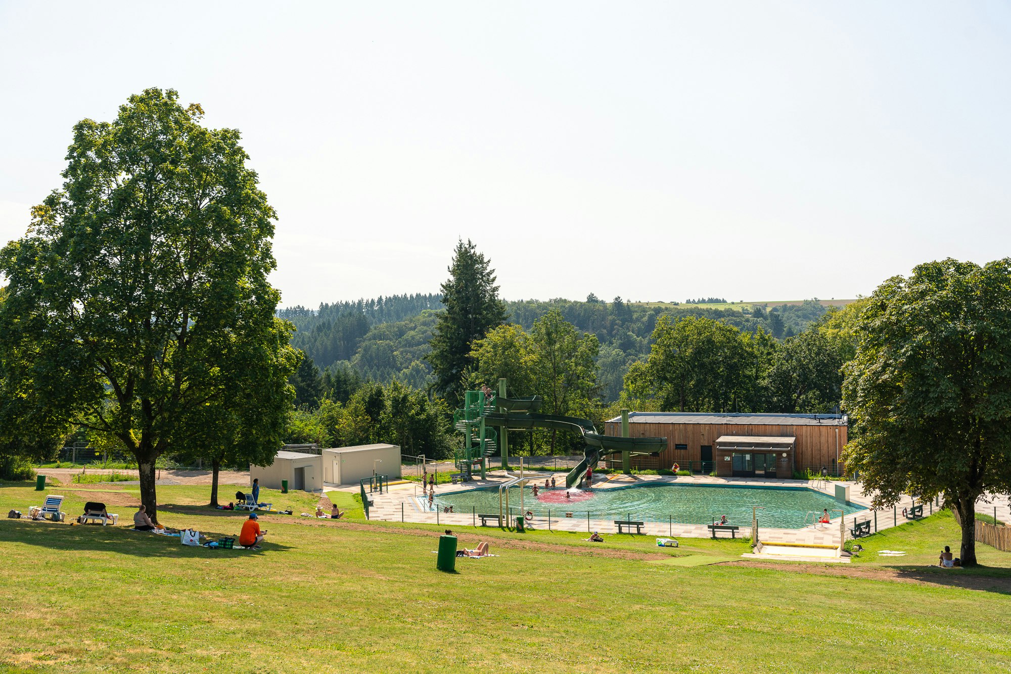 Schwimmbad auf dem Campingplatz mit Wasserrutsche, Liegewiese, Bäumen und Holzgebäude im Hintergrund.