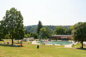 Schwimmbad auf dem Campingplatz mit Wasserrutsche, Liegewiese, Bäumen und Holzgebäude im Hintergrund.