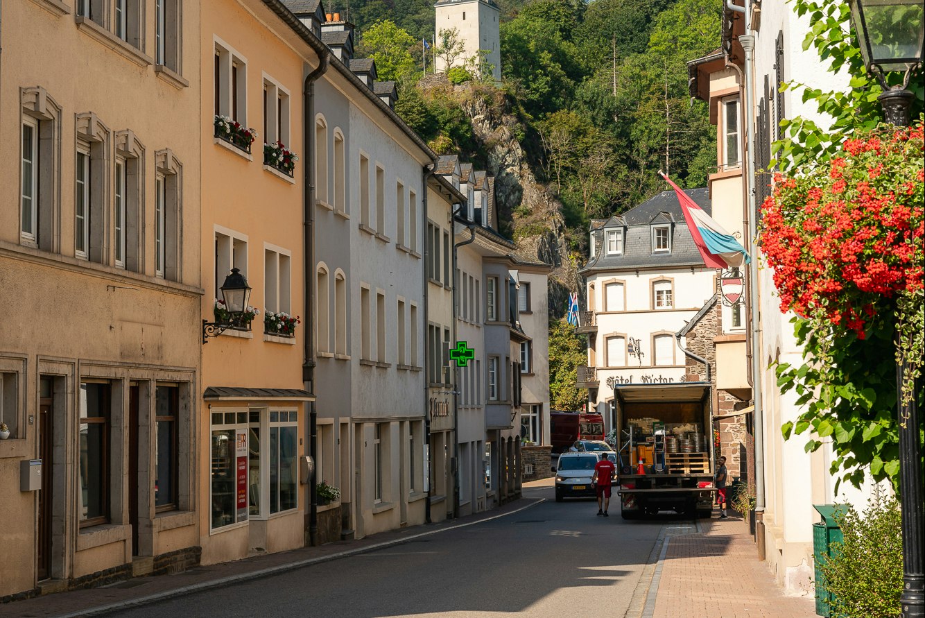 Altstadtgasse zwischen mehrstöckigen Gebäuden, Blumenampeln und einem Lieferwagen, im Hintergrund Wald und Felswand.