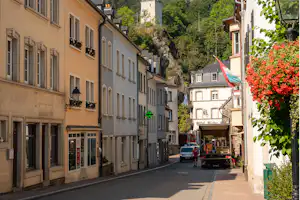 Altstadtgasse zwischen mehrstöckigen Gebäuden, Blumenampeln und einem Lieferwagen, im Hintergrund Wald und Felswand.