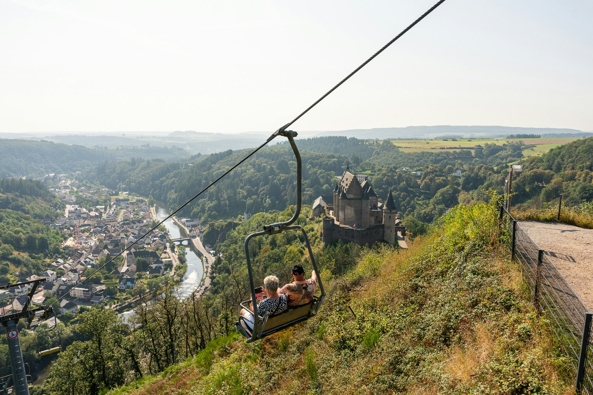 Seilbahn mit Sitzbank und Blick ins Tal: Fluss, Waldhügel und eine Burganlage im Vordergrund.