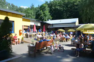 Camping-Terrasse mit vielen Gästen, Holzbänken und Tischen, gelben Sonnenschirmen vor einem Gebäude und im Hintergrund Bäume.