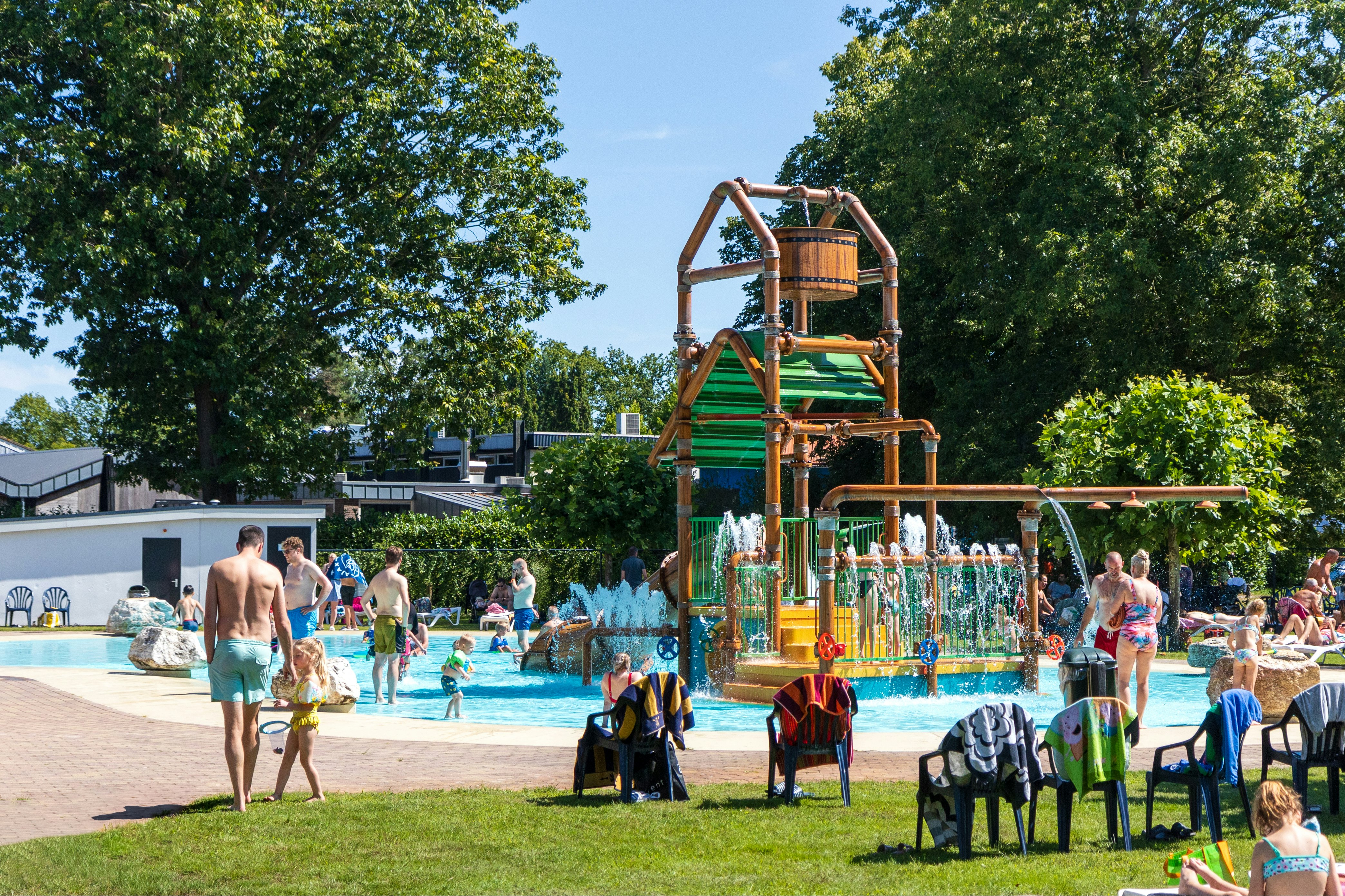 Wasserspiel im Schwimmbad mit grünem Rutschenturm, spritzendem Wasser und Familien am Beckenrand unter Bäumen.