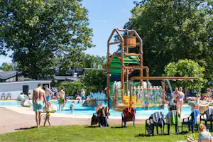 Wasserspiel im Schwimmbad mit grünem Rutschenturm, spritzendem Wasser und Familien am Beckenrand unter Bäumen.