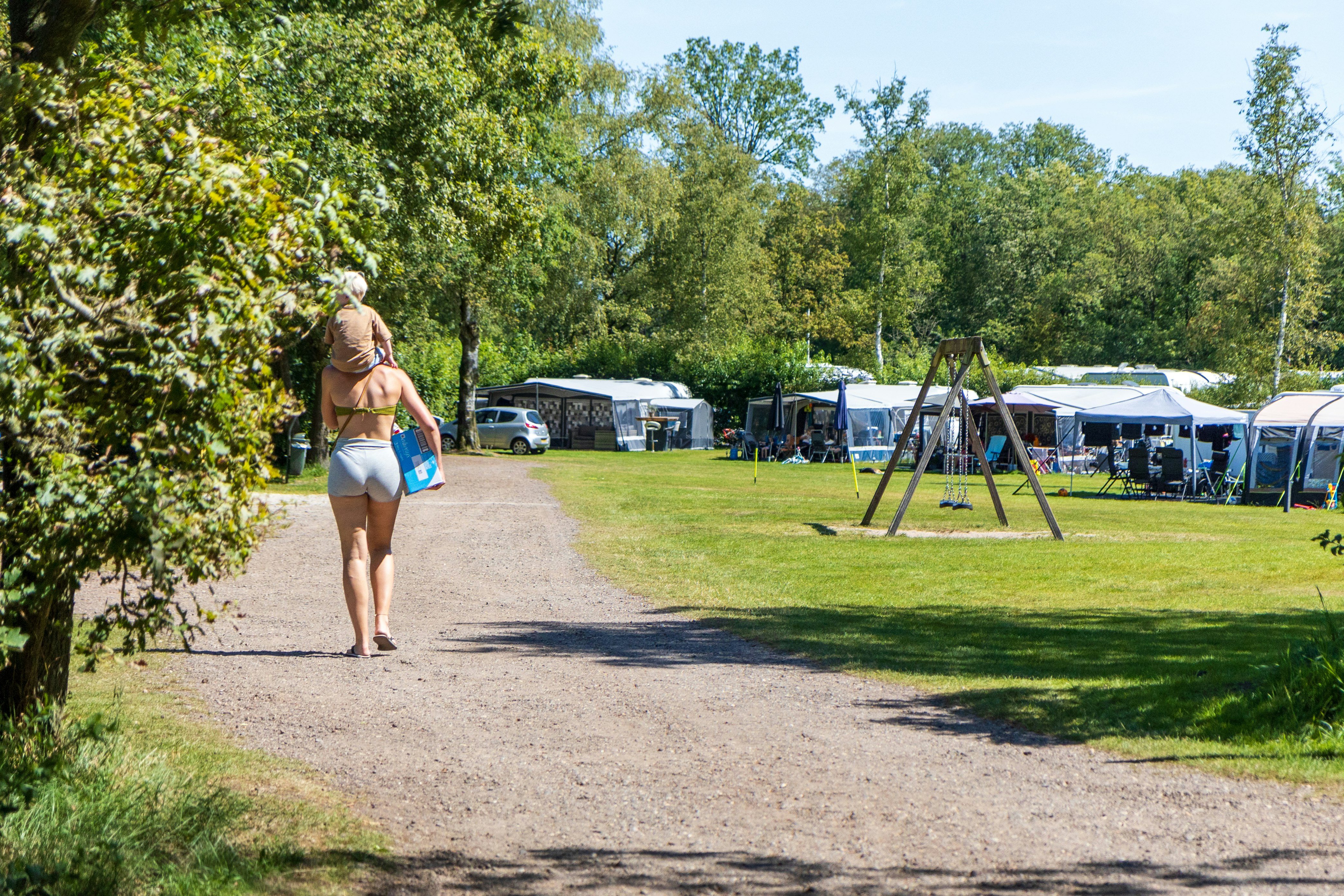 Person geht auf einem Kiesweg am Campingplatz vorbei, mit Wohnwagen, Zelten und Autos im Hintergrund unter Bäumen.
