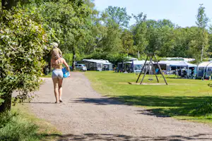 Person geht auf einem Kiesweg am Campingplatz vorbei, mit Wohnwagen, Zelten und Autos im Hintergrund unter Bäumen.