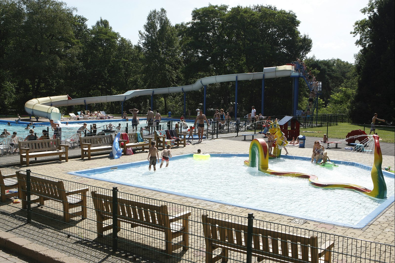 Schwimmbecken auf dem Campingplatz mit Wasserspiel, Liegen und Zaun, im Hintergrund Bäume und ein überdachter Bereich.