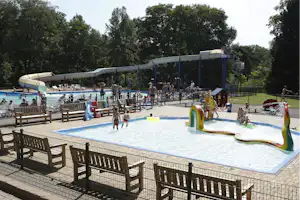 Schwimmbecken auf dem Campingplatz mit Wasserspiel, Liegen und Zaun, im Hintergrund Bäume und ein überdachter Bereich.