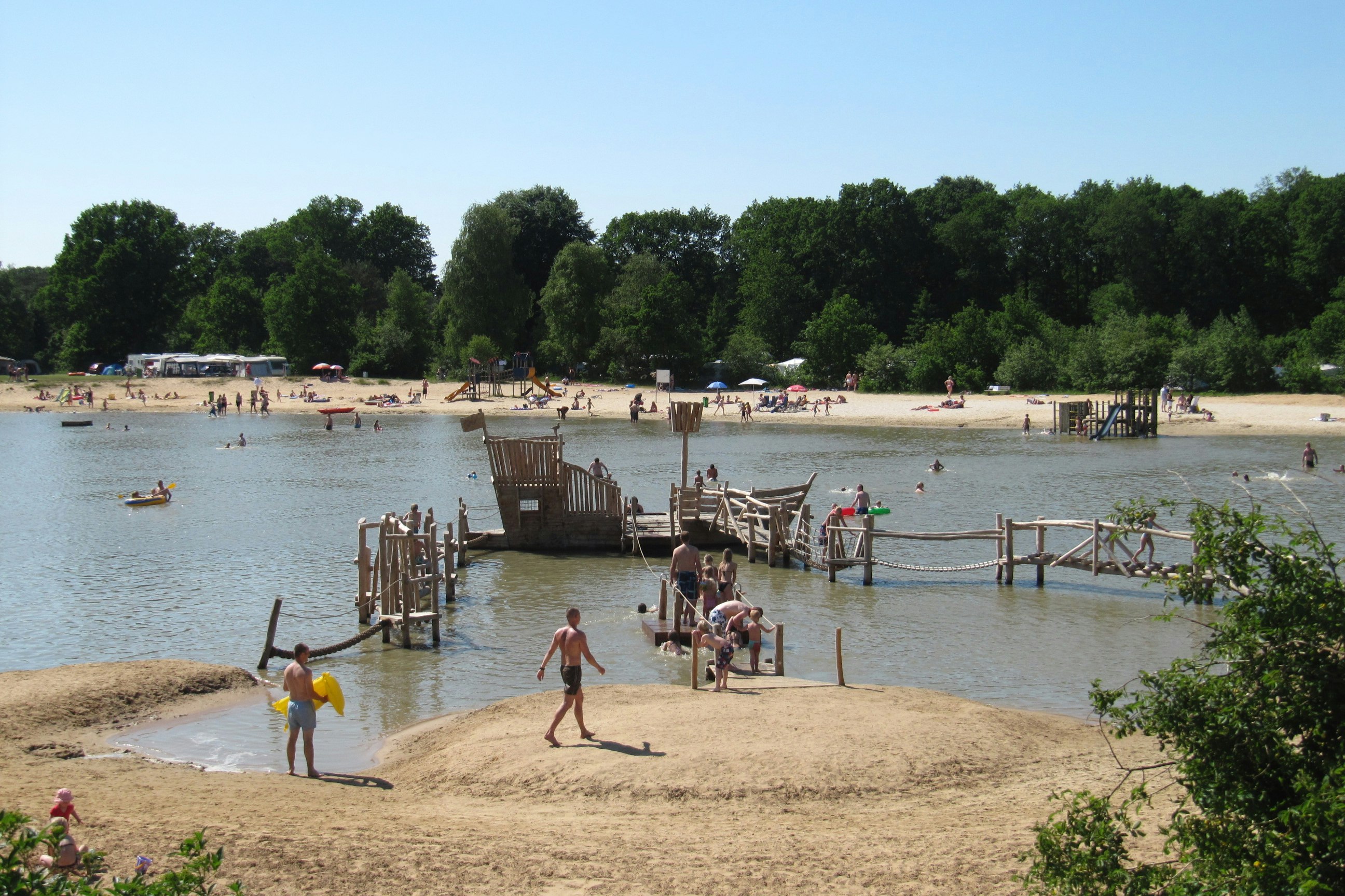 Blick auf den See mit Sandstrand und Stegen, Menschen am Ufer und im Wasser, Bäume am Rand.
